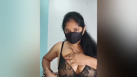 Media: A video shows a South Asian woman with long black hair, wearing a black lace bra and face mask, covering her breasts with her hand. The background features a light blue wall and a dark wooden door.