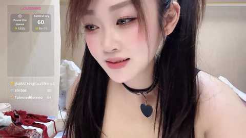 Video of an East Asian woman with long black hair, wearing a black choker with a heart pendant. She has light skin, pink blush, and subtle makeup. Background shows a messy bed with red and white bedding.