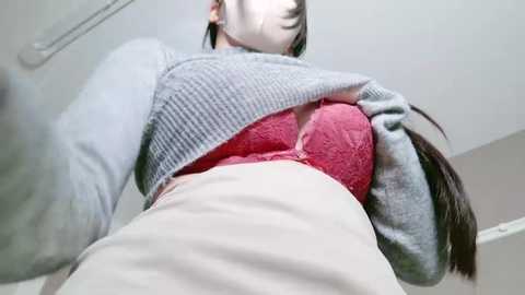 Video of a woman with fair skin and dark hair, wearing a grey sweater and a red lace bra, taken from a low angle emphasizing her chest.