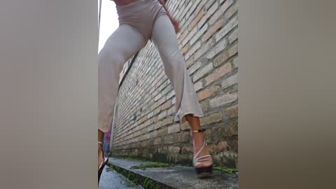 A video showing a person's legs in tight, light beige pants, walking on a wet, mossy sidewalk next to a weathered brick wall.
