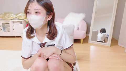Video of a young Asian woman in a white schoolgirl outfit, wearing a face mask, sitting on a white rug in a modern room with light wooden flooring and pastel furniture.