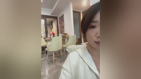 A video of an Asian woman with short brown hair, wearing a white hoodie, sitting in a modern, sparsely furnished room with light-colored chairs and a wooden table.