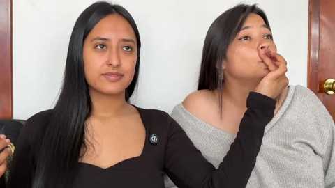 Video of two young South Asian women with medium skin tones and long black hair, one in a black top, the other in a gray off-shoulder sweater, both laughing.