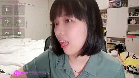 A video of an Asian woman with straight black hair and a bob haircut, wearing a green blouse, sitting on a bed with white sheets. Background shows a cluttered room with a white bookshelf and toys.