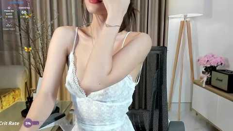Video of an Asian woman with light skin, wearing a white lace camisole, leaning forward, capturing a selfie in a modern room with beige curtains, a black chair, and a pink flower arrangement.