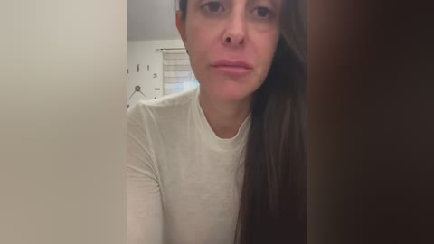 Video of a young woman with long brown hair, wearing a white long-sleeve shirt, in a dimly lit room with a white wall and a window with blinds.