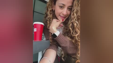 Video of a smiling, curly-haired woman with light brown skin, wearing a brown sweater, holding a red Starbucks cup, sitting indoors with a blurred background.
