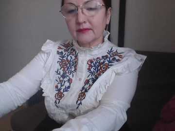 Video of a middle-aged woman with glasses, fair skin, and brown hair, wearing a white lace-up blouse with intricate embroidery. She sits against a dark background, looking down with a neutral expression.
