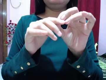 Video of a woman with long black hair, wearing a teal long-sleeve shirt with gold buttons, adjusting her earrings. Background includes a red curtain, a wooden door, and a floral arrangement.