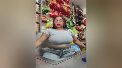 Video of a young Asian woman with glasses and light skin, wearing a tight gray crop top and matching pants, sitting behind a cash register in a brightly lit convenience store.