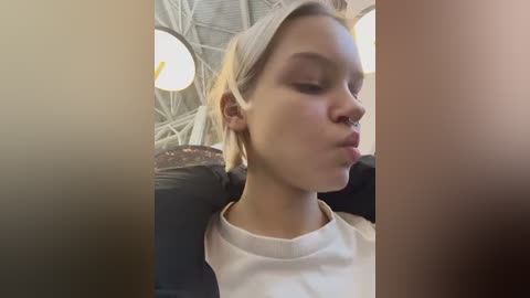 Video of a young woman with blonde hair, pale skin, and a septum piercing, wearing a white T-shirt, sitting in a dark interior with circular light fixtures and a metallic ceiling.