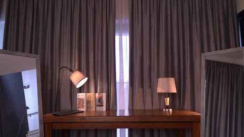 Video of a cozy bedroom with a wooden table holding two table lamps, framed photos, and a wall with grey curtains.