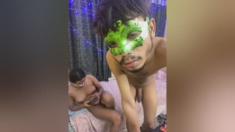 Video of a naked Asian man with a green Venetian mask, leaning forward on a bed with a pink quilt, beside a nude woman with a black mask, in a dimly lit room with blue string lights.