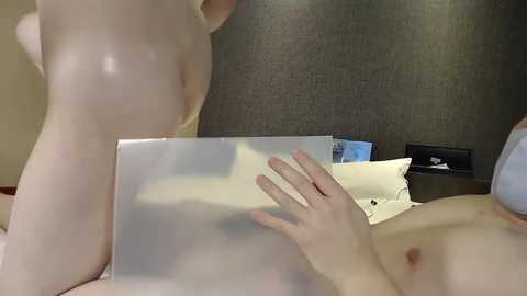 A video of a nude woman with pale skin and a slender physique, partially obscured by a translucent shower curtain, sitting on a bed with a dark grey headboard.