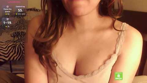 Video of a fair-skinned woman with wavy brown hair, wearing a beige lace-trimmed camisole, showing cleavage, in a cozy, dimly lit bedroom with a zebra-print blanket and a digital timer display.