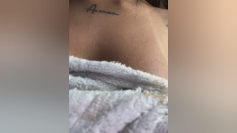 Video of a woman's upper chest and neck, showing a tattoo of the word \"Amor\" in cursive script, partially covered by a soft, light blue towel.