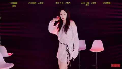 Video of a woman with long dark hair, wearing a white, off-the-shoulder crochet dress, singing into a microphone, surrounded by modern white chairs against a dark backdrop, with stage lighting.