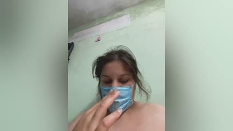 A video of a woman with light brown skin, dark hair, and a blue surgical mask, covering her nose and mouth, standing in a small room with light green walls and a white ceiling.