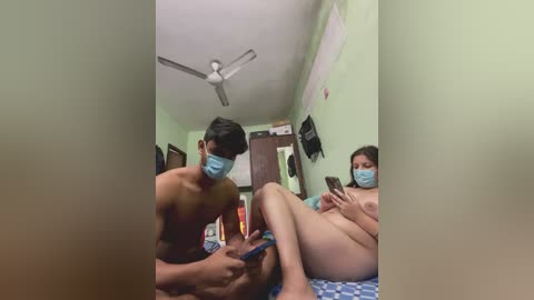 Video of two nude Asian individuals, a man and a woman, in a dimly lit bedroom. The man, wearing a blue face mask, is massaging the woman's leg.