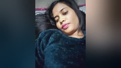 Video of a young woman with medium brown skin and straight black hair, lying on her side, wearing a dark blue sweater, with a serene expression and closed eyes, set against a blurred background.