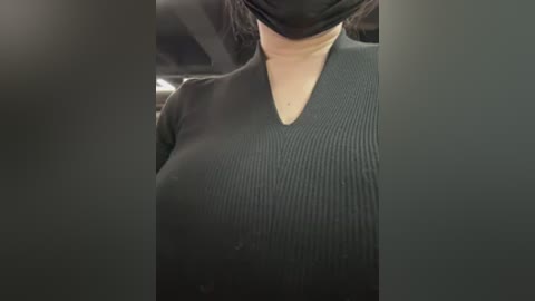 A close-up video of a person wearing a black face mask and a black, ribbed V-neck sweater. The background shows a car interior with seat belts visible.