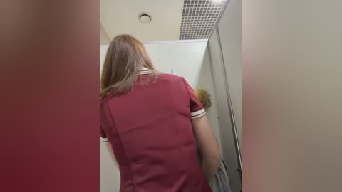 Media: A video shows a person with shoulder-length light brown hair in a red uniform with white trim, walking away in a narrow hallway with beige walls and a tiled ceiling.