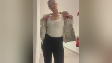 Video of a slender, light-skinned woman with glasses, wearing a white tank top and high-waisted black leggings, standing in a hallway with beige walls and a red fire extinguisher on the right.
