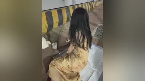 Video of a woman in a mustard-yellow dress with floral patterns, seated on a white cushioned bench. Background features a yellow and black striped barrier, a cobblestone pavement, and a white building.