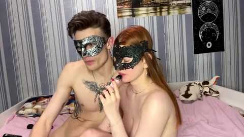 Video of a nude, fair-skinned man with a tattooed chest and short brown hair, wearing a black masquerade mask, kissing a red-haired, fair-skinned woman with a black lace mask. They sit on a bed with striped wallpaper and a cat.