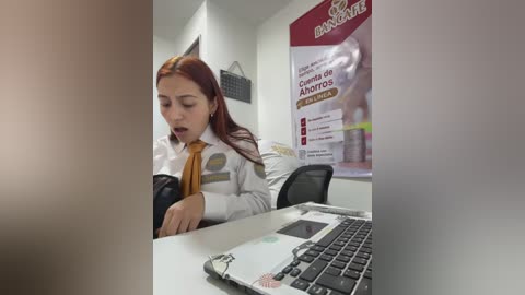 Video of a young woman with long red hair, wearing a white shirt and yellow tie, looking surprised, sitting at a desk in a modern office with a laptop in front of her.