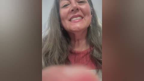 Video of a middle-aged woman with long, wavy, gray hair, wearing a red sweater, smiling broadly. The background is blurred, with a focus on her face.
