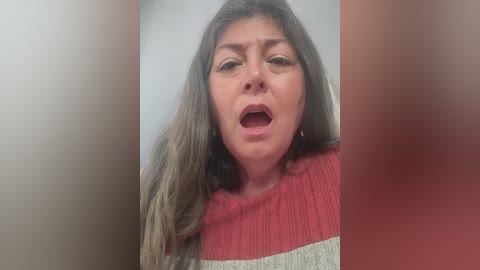 A video of a woman with long, straight brown hair, wearing a red and beige striped sweater, mouth open in an expression of surprise or shock, with blurred background.
