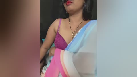 Video of a woman with medium brown skin, wearing a purple bra and light blue saree, standing in front of a blurred background. She has long, dark hair and wears red lipstick.