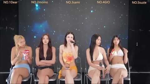 Video of five women seated on chairs, each holding a microphone, wearing various colorful outfits against a dark background with starry effects.