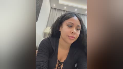 Video of a young Black woman with medium skin tone, long black hair, wearing a black lace-up top, standing in a modern room with grey curtains and recessed lighting.