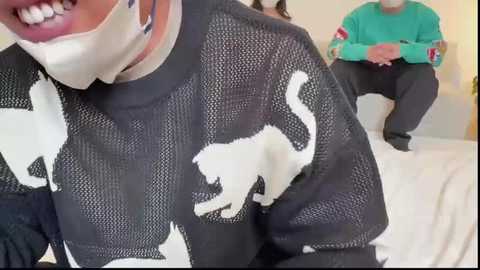 Video of a woman in a black sweater with white cat silhouette, grinning with white mask covering mouth, sitting on a white couch next to a man in a green sweater and black pants.