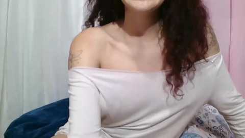 Video of a young woman with curly dark hair, light skin, wearing a white off-shoulder top, and a tattoo on her left shoulder. Background includes blue and white curtains, and pastel pink wall.