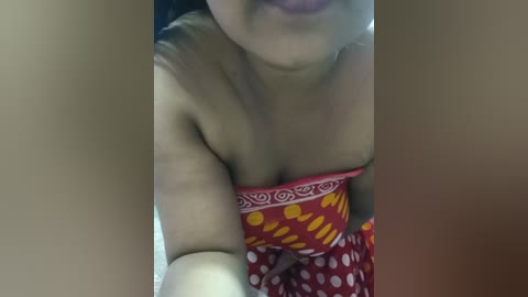 A video of a light-skinned woman with a medium build, wearing a red and yellow patterned strapless dress, partially covered by her arm, which is raised. Her face is partially obscured. The background is blurred, suggesting an indoor setting.