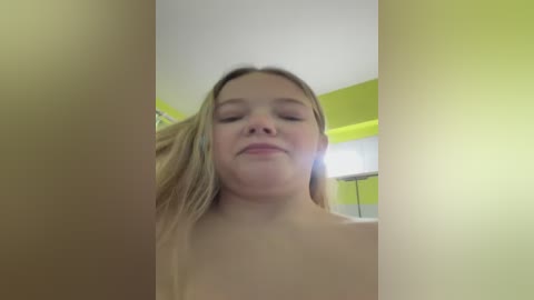 A video of a topless young woman with fair skin, blonde hair, and closed eyes, standing in a bright yellow room with a white ceiling. The image is slightly blurred.