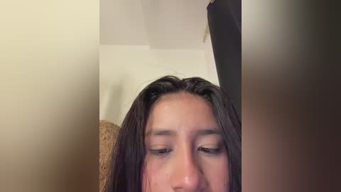 Video of a young woman with long, dark hair, looking down, in a dimly lit room with beige walls, a textured light brown couch, and a black curtain in the background.