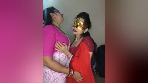 Video of two South Asian women, one in a pink blouse, the other in a red sari, wearing gold masks, embracing, in a dimly lit room.