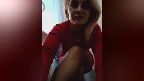 Video of a blonde woman with dark makeup, wearing a red top, crouching in a dimly lit room with red curtains.