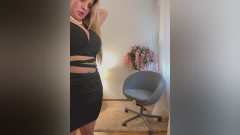 A video of a curvaceous woman in a tight black dress with cutouts, standing in a minimalist room with a gray chair and pink floral arrangement.