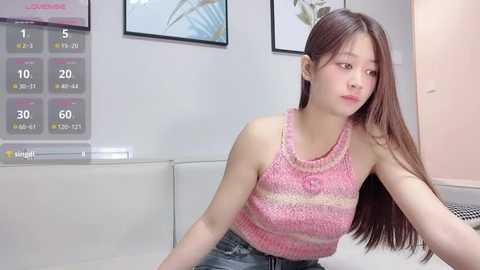 Video of an Asian woman with long brown hair, wearing a pink-striped tank top, sitting indoors against a white wall with a calendar and framed art.