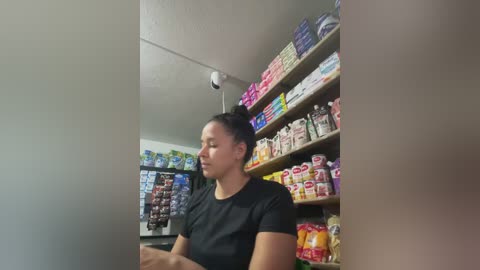 Video of a Latina woman with dark hair tied back, wearing a black t-shirt, standing in a convenience store with brightly lit shelves of snacks and beverages.