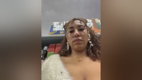 Video of a Black woman with curly hair, wearing a white lace dress, sitting in a grocery store. She has a neutral expression, surrounded by colorful products on shelves.