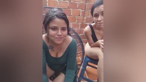 Video of a woman with dark hair, wearing a green top, sitting on a wicker chair beside another woman, both looking towards the camera in a brick-walled outdoor setting.