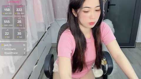 Video of a young Asian woman with long black hair and fair skin, wearing a pink T-shirt, sitting at a desk. She has a neutral expression. Background includes a glass wall, a white curtain, and a digital display showing temperature readings.