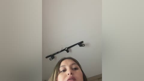 Video of a young woman with fair skin, light brown hair, and a neutral expression, looking upwards. She is partially visible from the neck down. A black ceiling fan is mounted on a white ceiling.