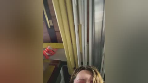 A video captures a partially obscured, light-skinned person with shoulder-length blonde hair standing in a dimly lit, narrow hallway with yellow and grey curtains.